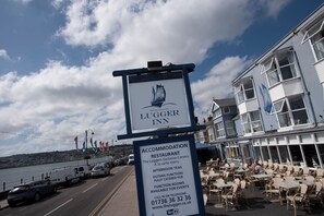 Une enseigne indiquant l’emplacement du Lugger Inn, un établissement hôtelier et un restaurant situés en bord de mer, avec une vue sur la mer et des voitures garées en arrière-plan.