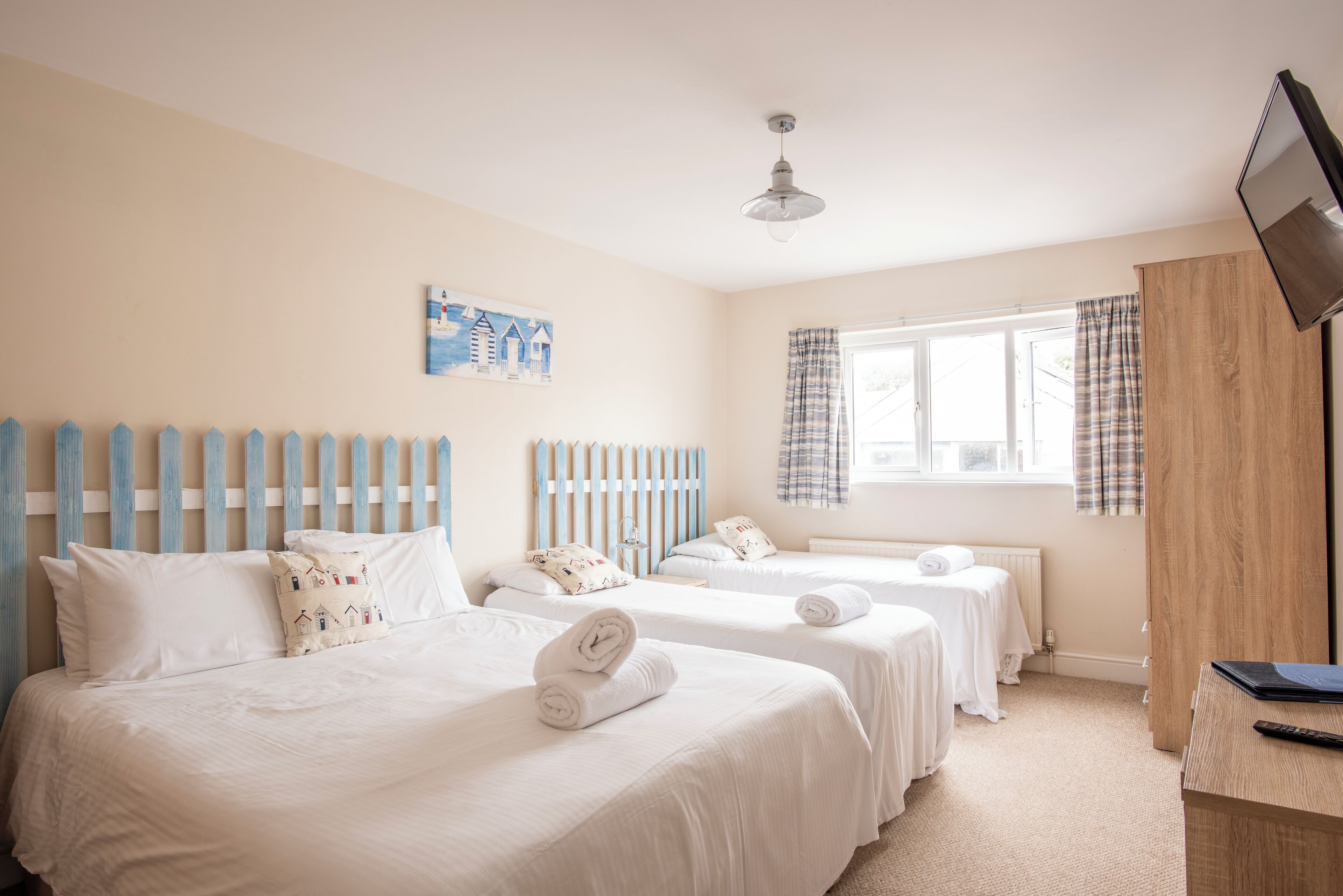 A hotel room with two beds, a wooden headboard, a window with curtains, a flat-screen TV, and a painting on the wall.