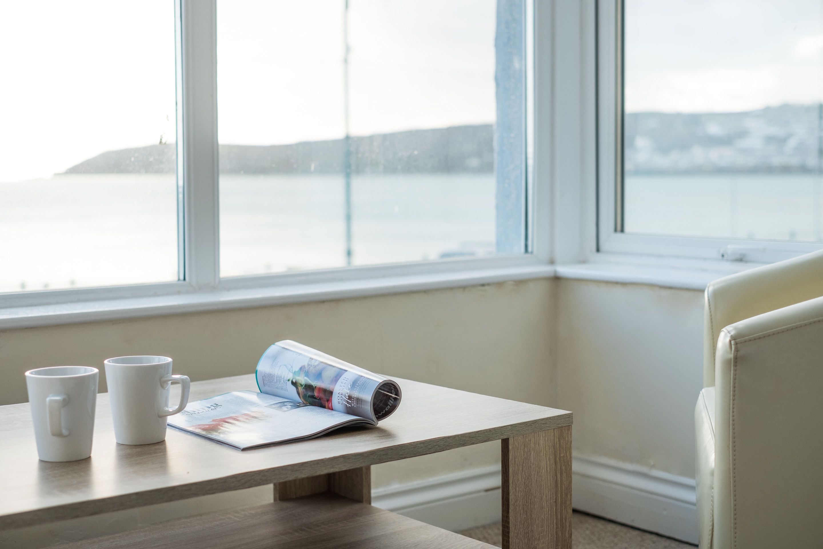 Una mesa con dos tazas blancas y una revista, una ventana que da a una masa de agua y una tierra distante.