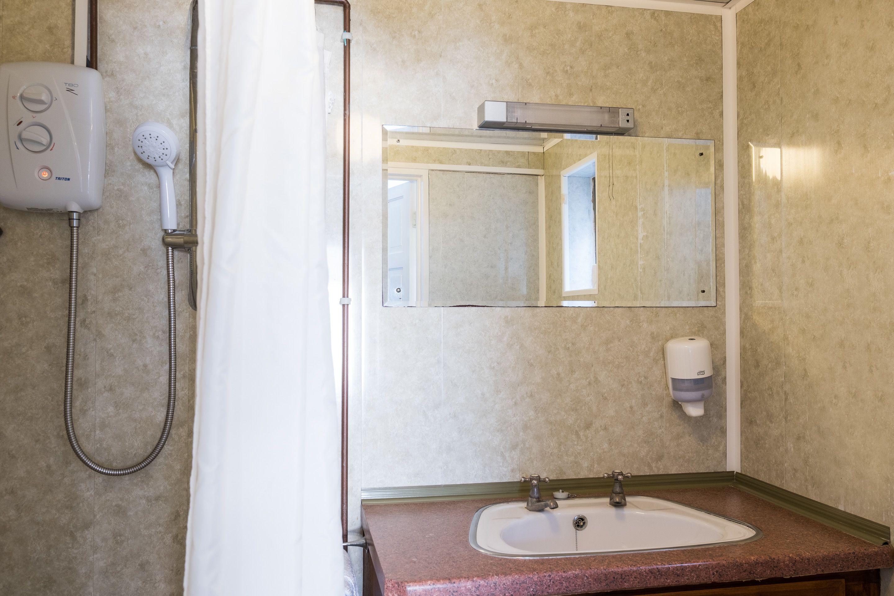 A bathroom with a shower, a sink, a mirror, and a soap dispenser.