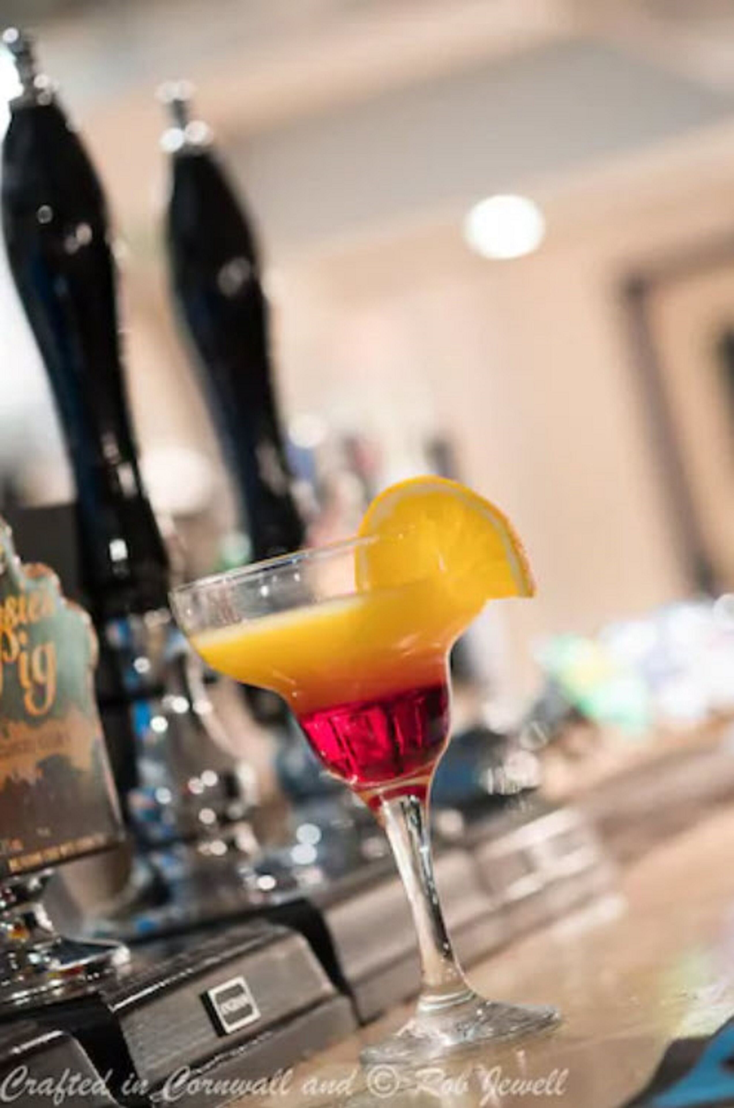 Cocktail-lasi, jonka reunalla on appelsiiniviipale, on asetettu baaritiskin päälle, ja taustalla näkyy oluthanat.