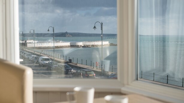 A view from a window overlooking a coastal road with parked cars, a pedestrian walkway, and a distant castle-like structure. - The Lugger Inn (Penzance)