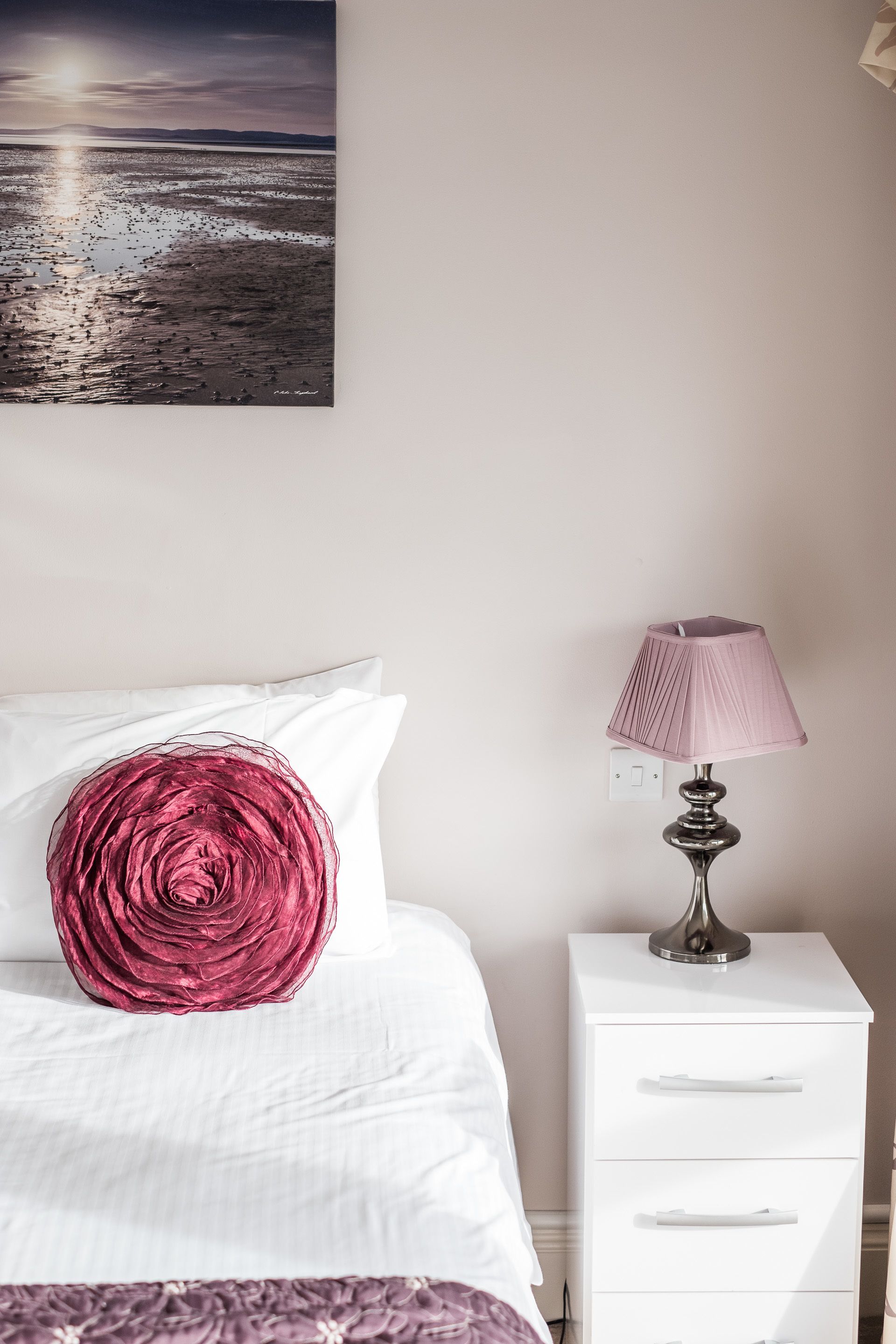 Ein Schlafzimmer mit einem Bett, einem Nachttisch mit Lampe, einem weißen Beistelltisch und einem Wandbild, das einen Sonnenuntergang über Wasser zeigt.