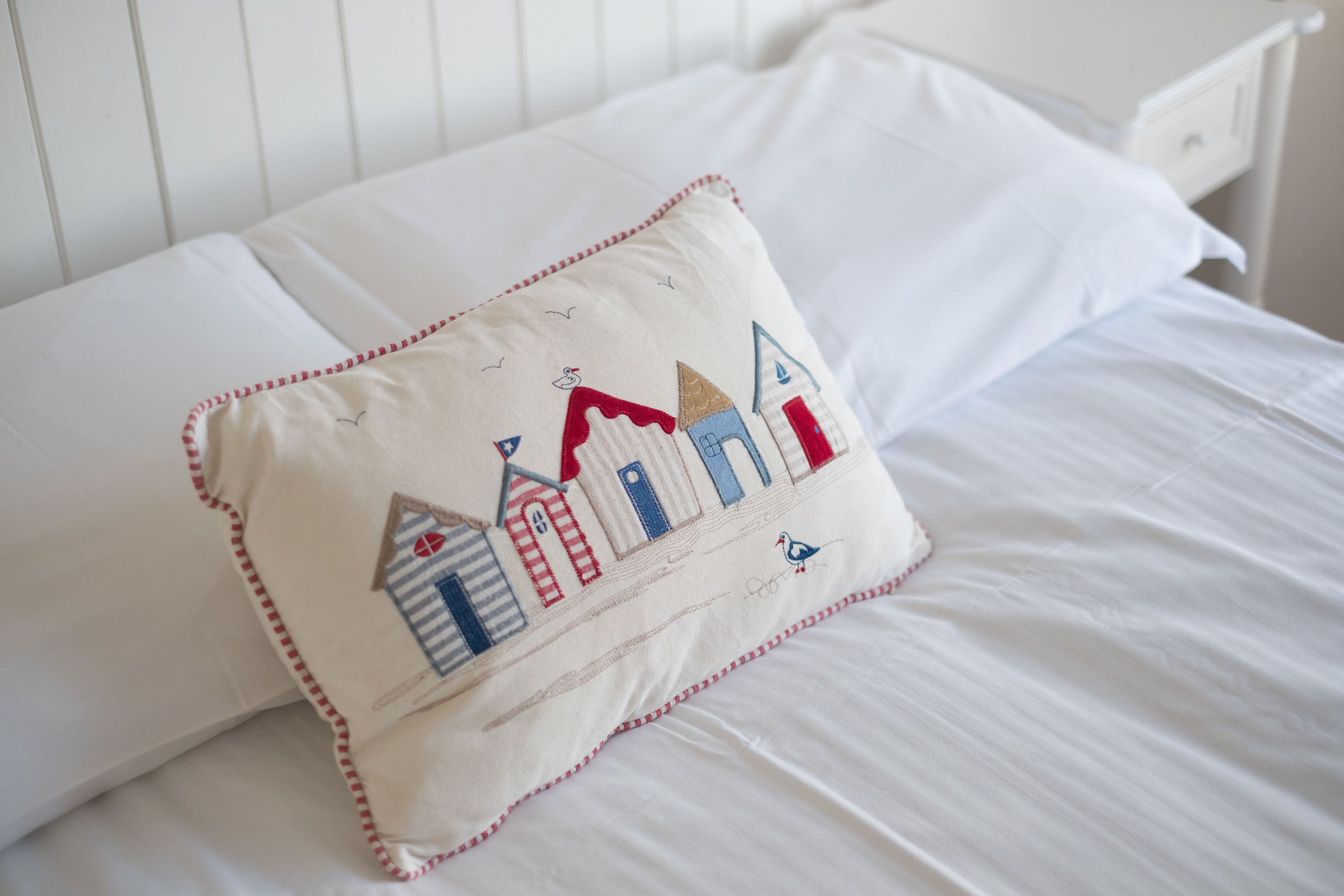 Una cama con sábanas blancas y un almohadón con diseño bordado de edificios y un pájaro.