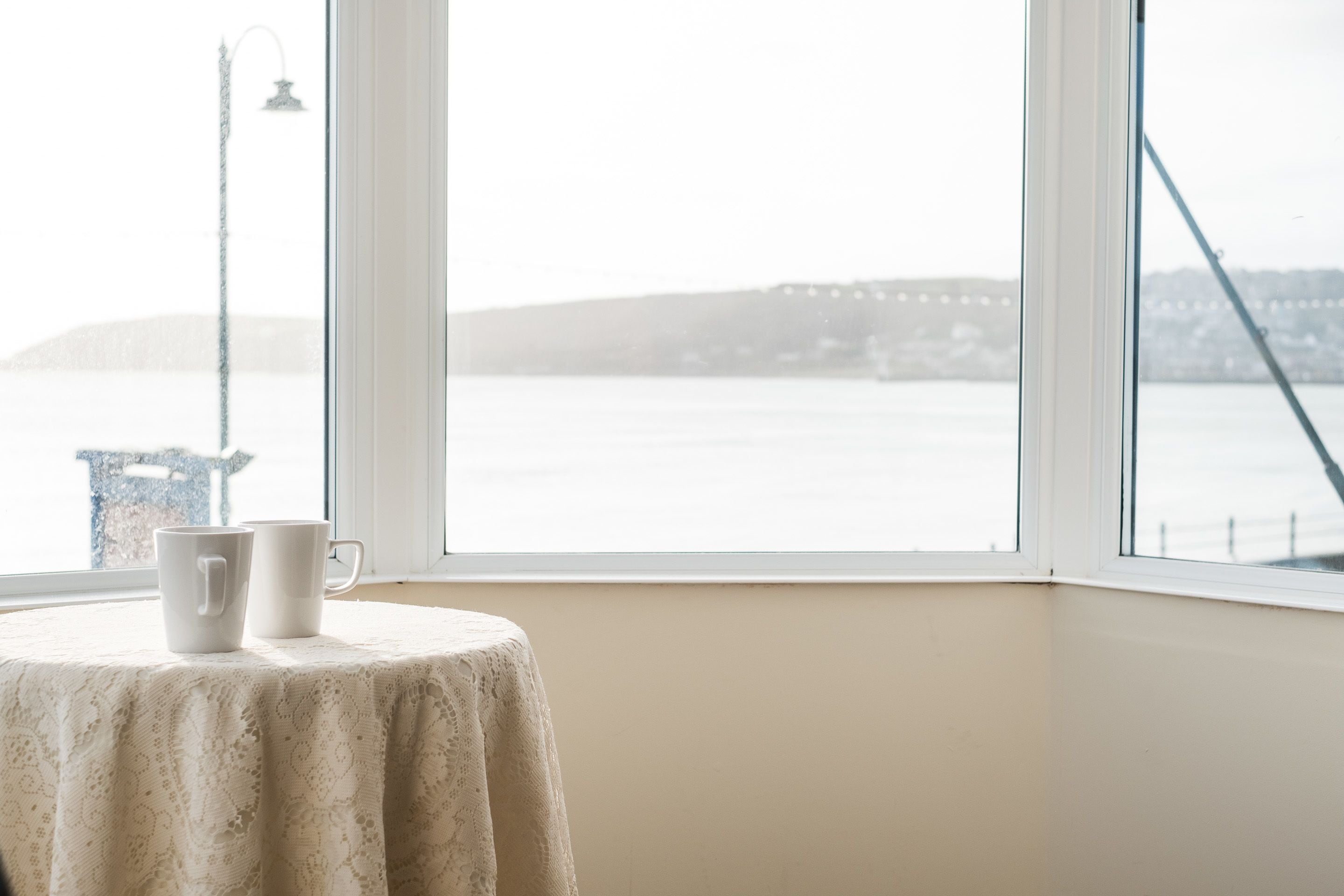 Une table recouverte d’une nappe en dentelle blanche, avec deux tasses blanches, placée près d’une fenêtre donnant sur un pont et sur l’eau.