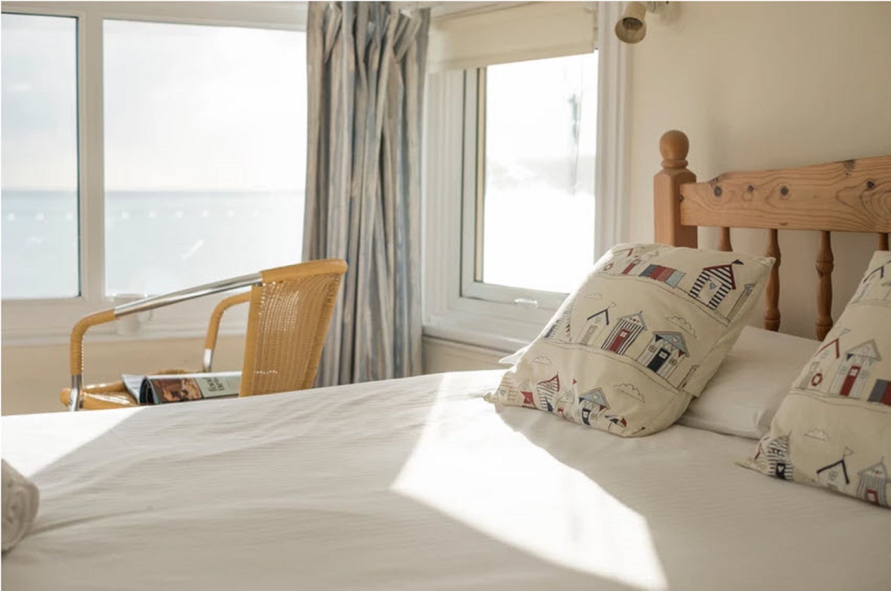 Een slaapkamer met een bed, een houten hoofdbord, een rieten stoel en uitzicht op zee door grote ramen.
