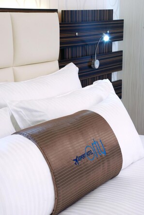 Une chambre d’hôtel avec un lit, du linge de lit blanc, un coussin décoratif marron et une tête de lit dotée d’une lampe intégrée.