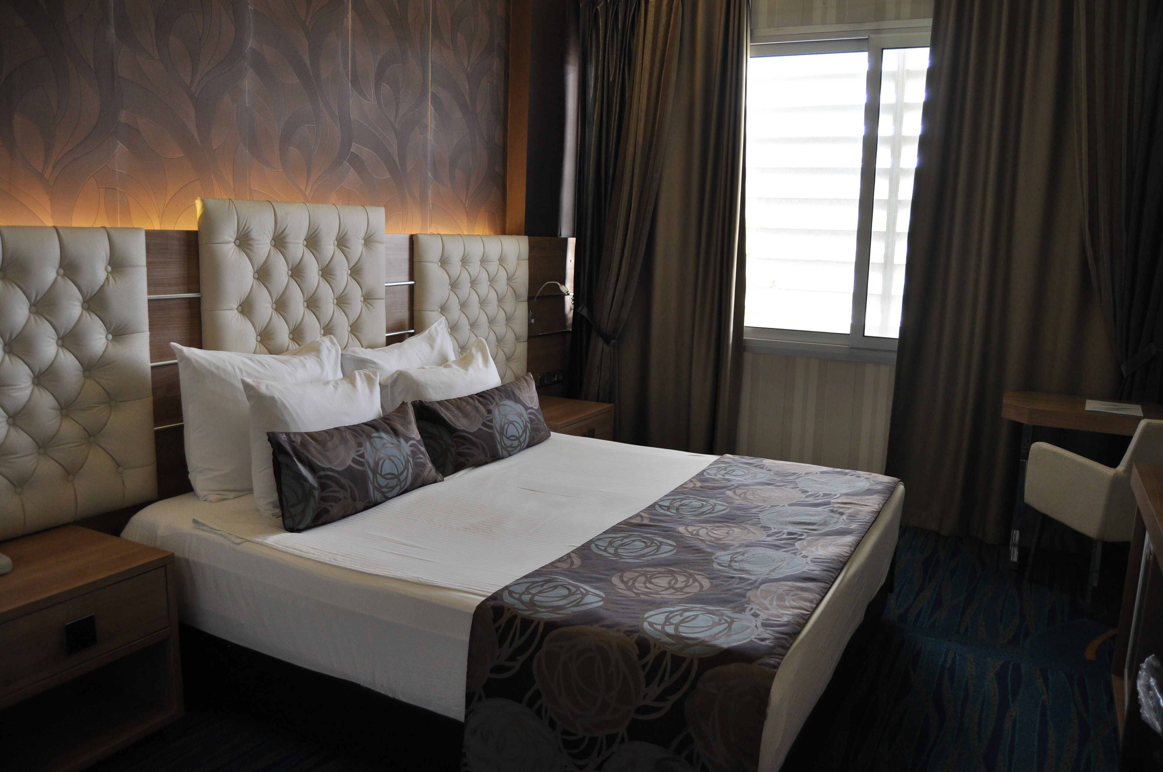 A hotel room with a large bed, a wooden nightstand, a desk, and a window with curtains.