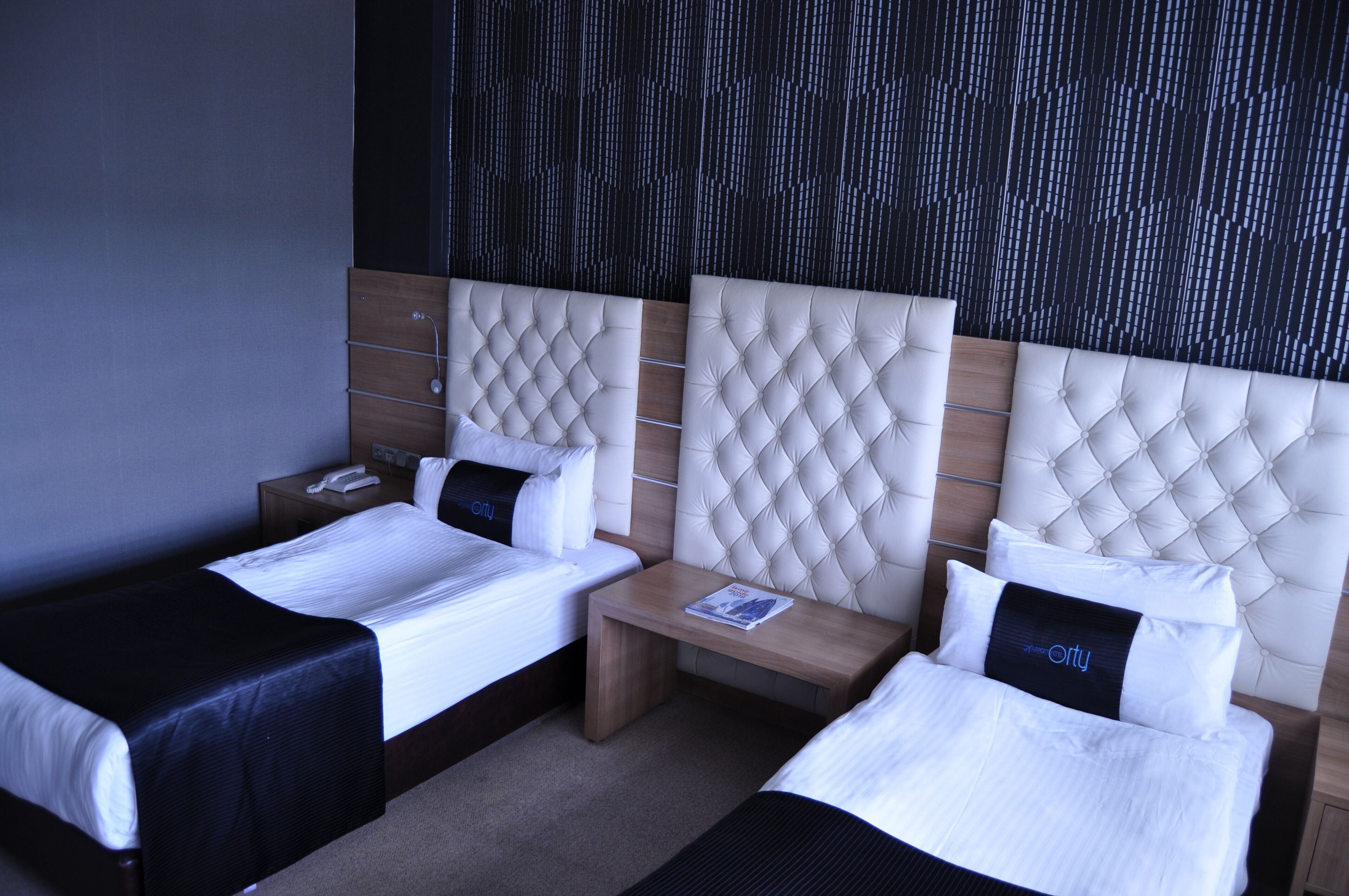 Ein Hotelzimmer mit zwei Betten, einem hölzernen Kopfteil, einem kleinen Nachttisch mit Telefon und einer Wand mit Struktur.