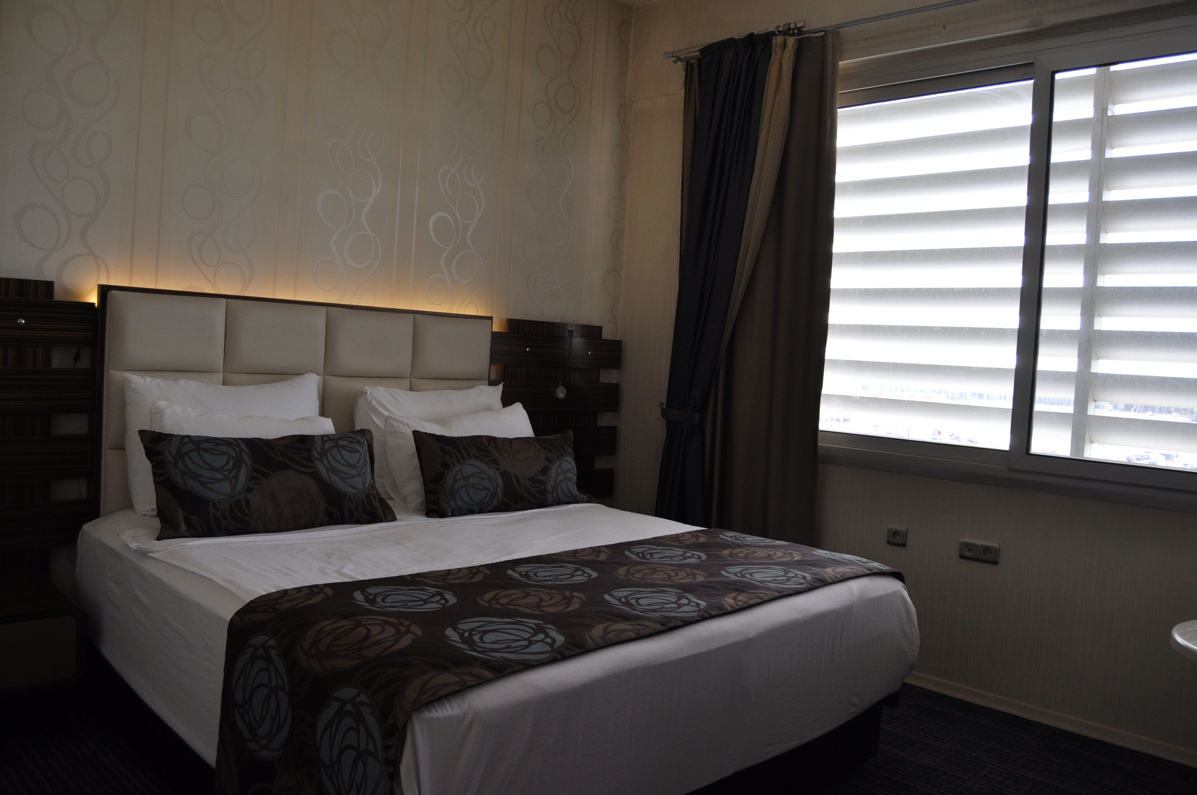 A hotel room with a large bed, a headboard, a window with blinds, and a nightstand.