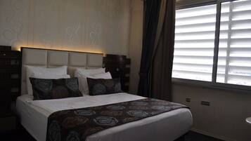 A hotel room with a large bed, a headboard, a window with blinds, and a nightstand.