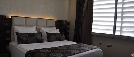 A hotel room with a large bed, a headboard, a window with blinds, and a nightstand.