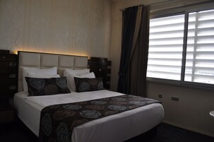 A hotel room with a large bed, a headboard, a window with blinds, and a nightstand.