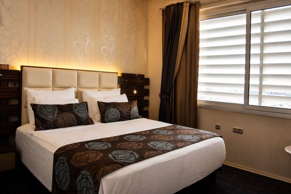 Een hotelkamer met een bed, een hoofdbord, een raam met jaloezieën en een nachtkastje.