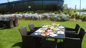 Un área de comedor al aire libre con mesas dispuestas para el desayuno, rodeada de un jardín con plantas en flor y un edificio grande al fondo.