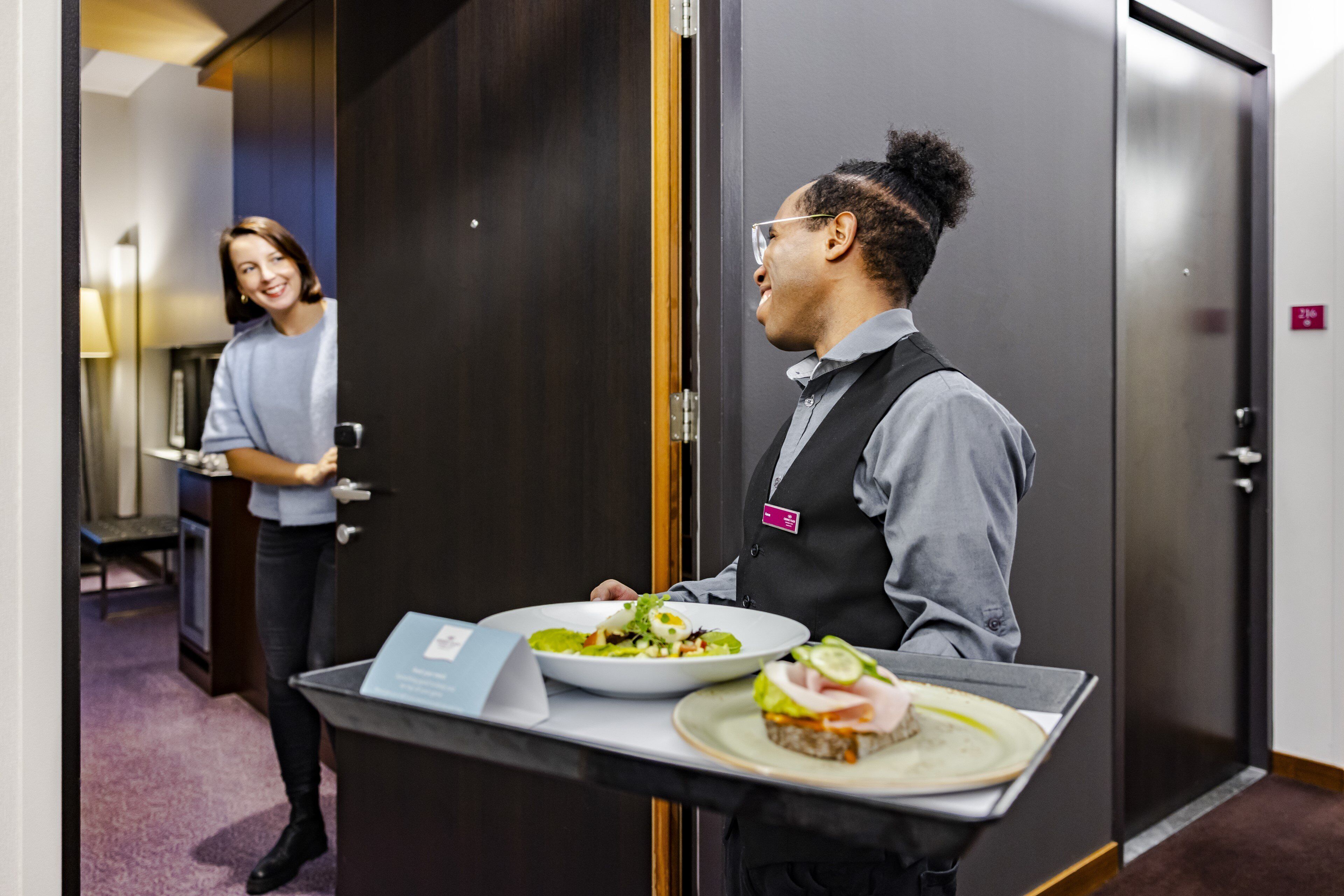 Una hall d'albergo con una reception, un ospite che porta un vassoio con del cibo, e una porta sullo sfondo.