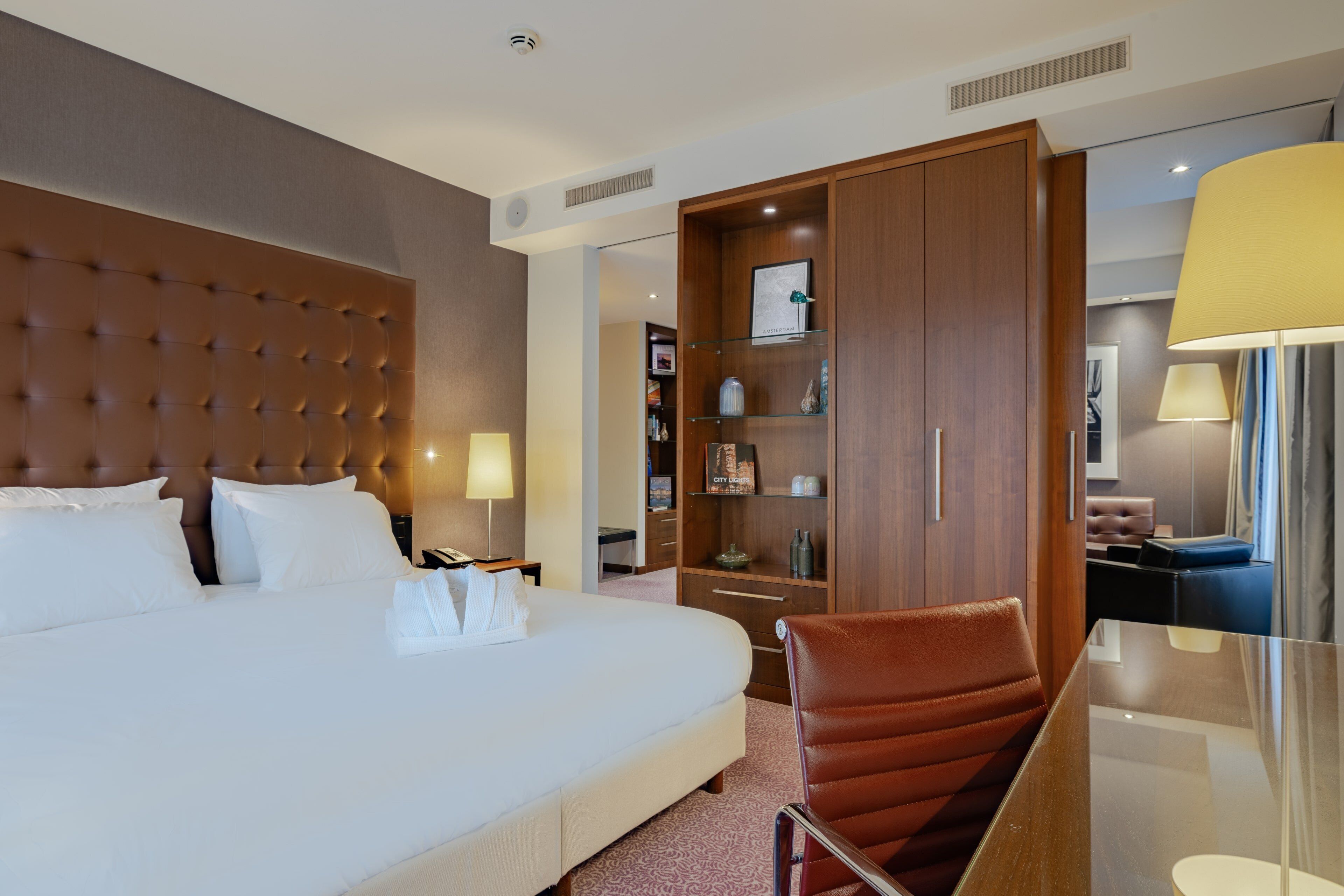 Een hotelkamer met een groot bed, een houten garderobe, een zithoek met een stoel en een bureau met een lamp.