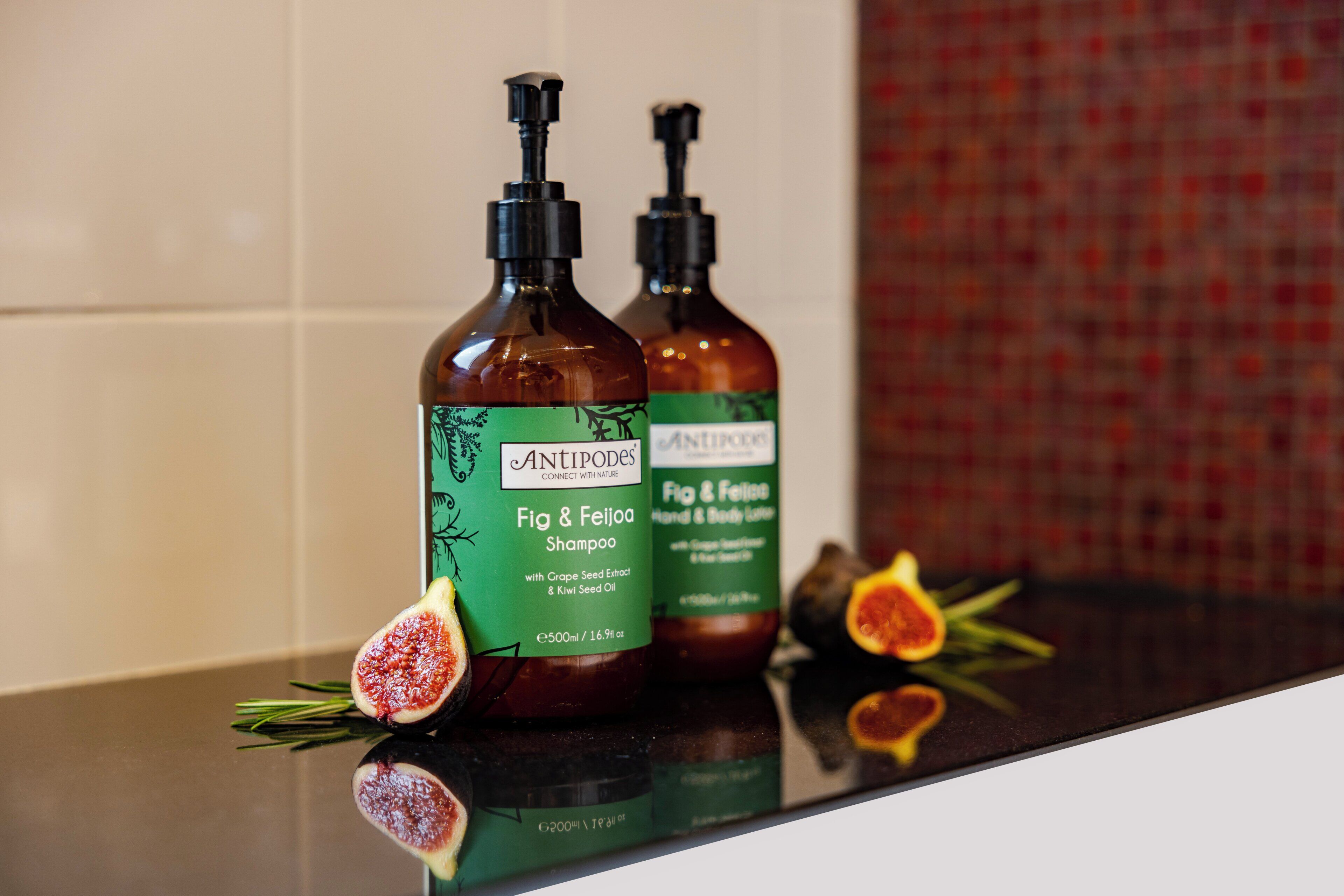 Dos botellas de champú Antipodes Fig & Feijoa sobre una encimera reflectante, adornadas con higos y romero.