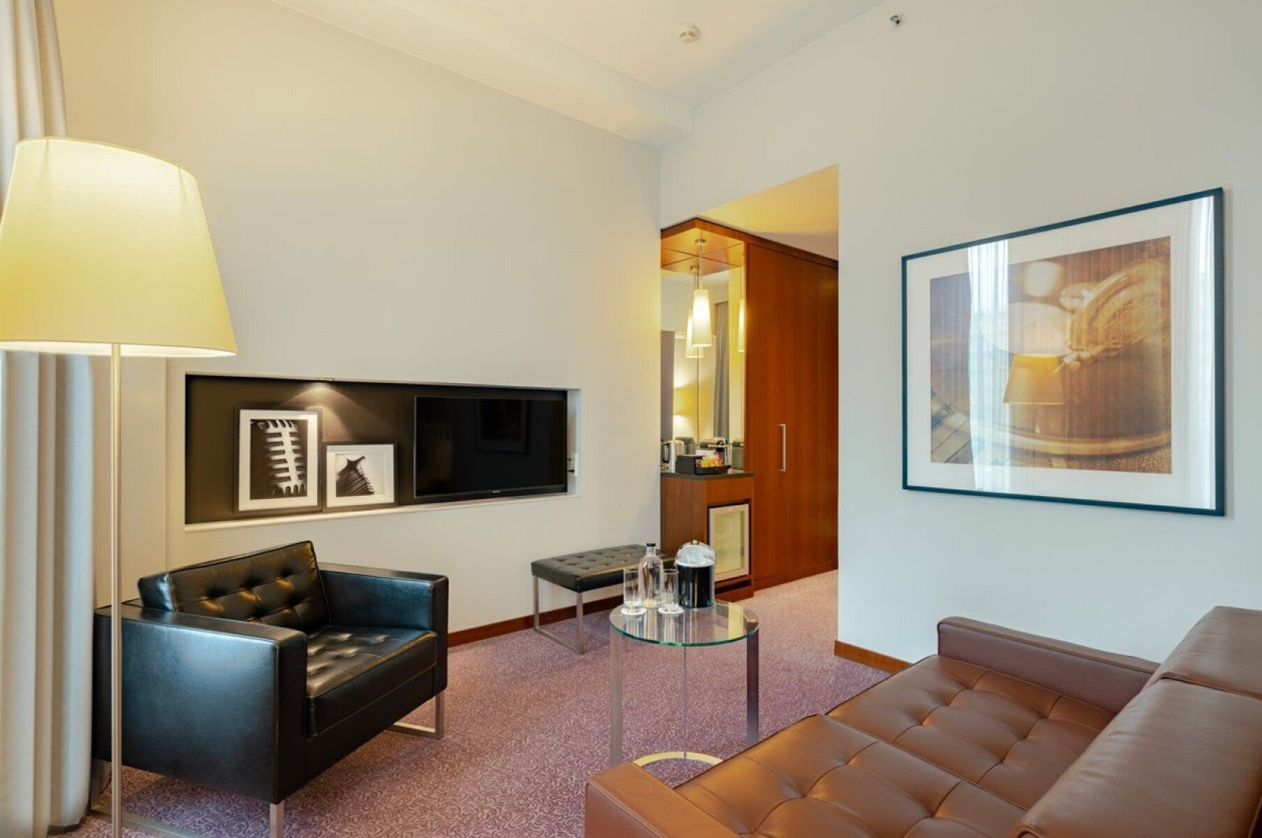 Ein modernes Hotelzimmer mit einer Ledersofa, einem Glastisch, einem flachbildfernseher an der Wand, einem gerahmten Kunstwerk und einer Lampe.