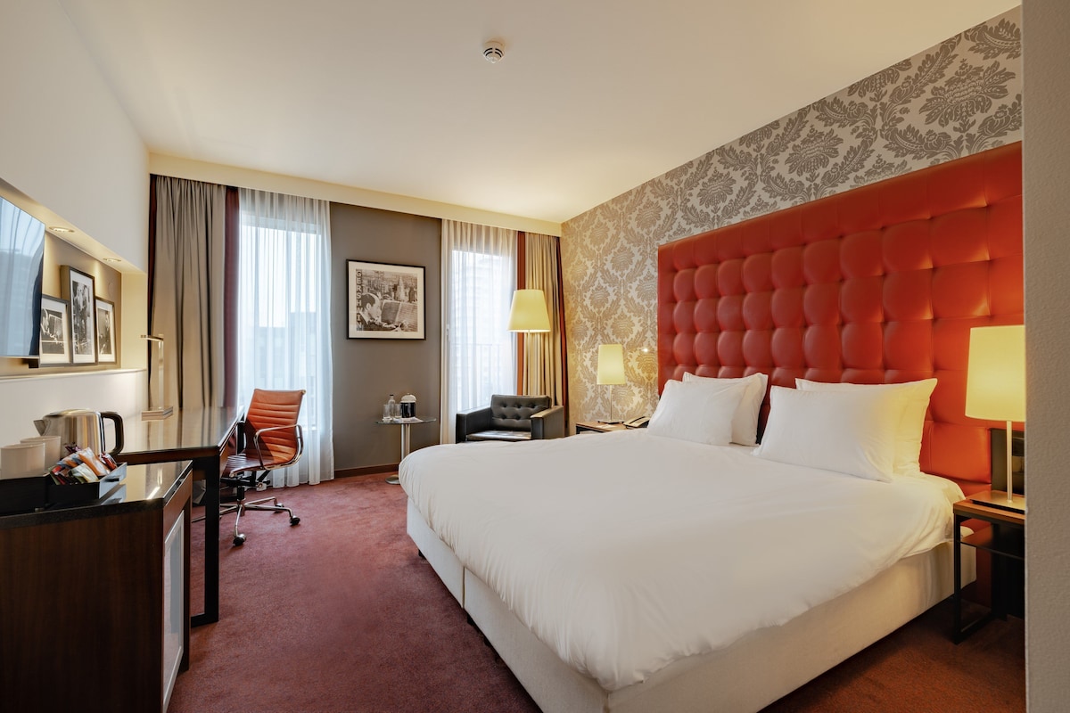 A hotel room with a large bed, a desk with a chair, a small table with a lamp, and a window with curtains.