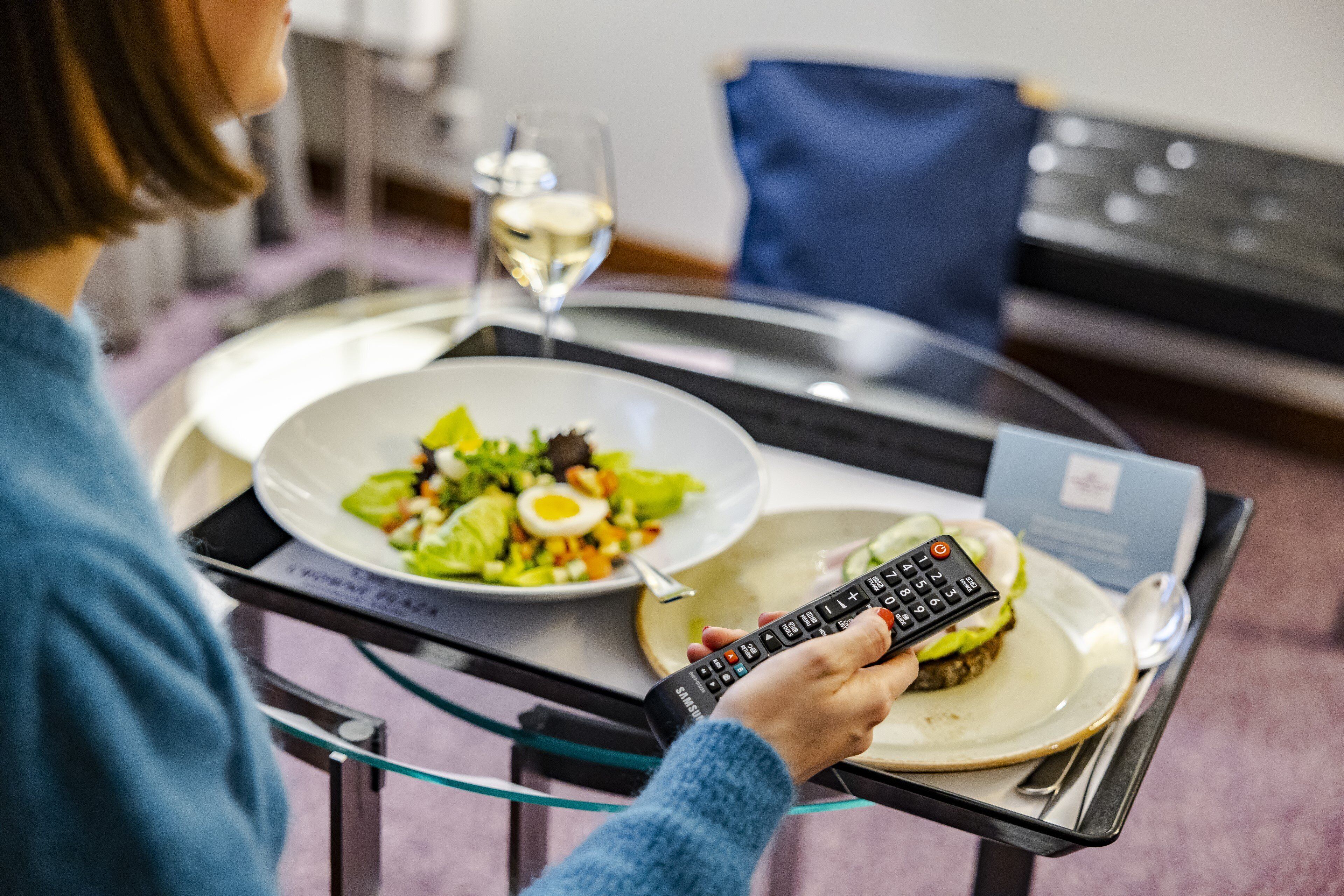 A person holding a remote control, sitting at a table with a plate of food and a glass of wine.