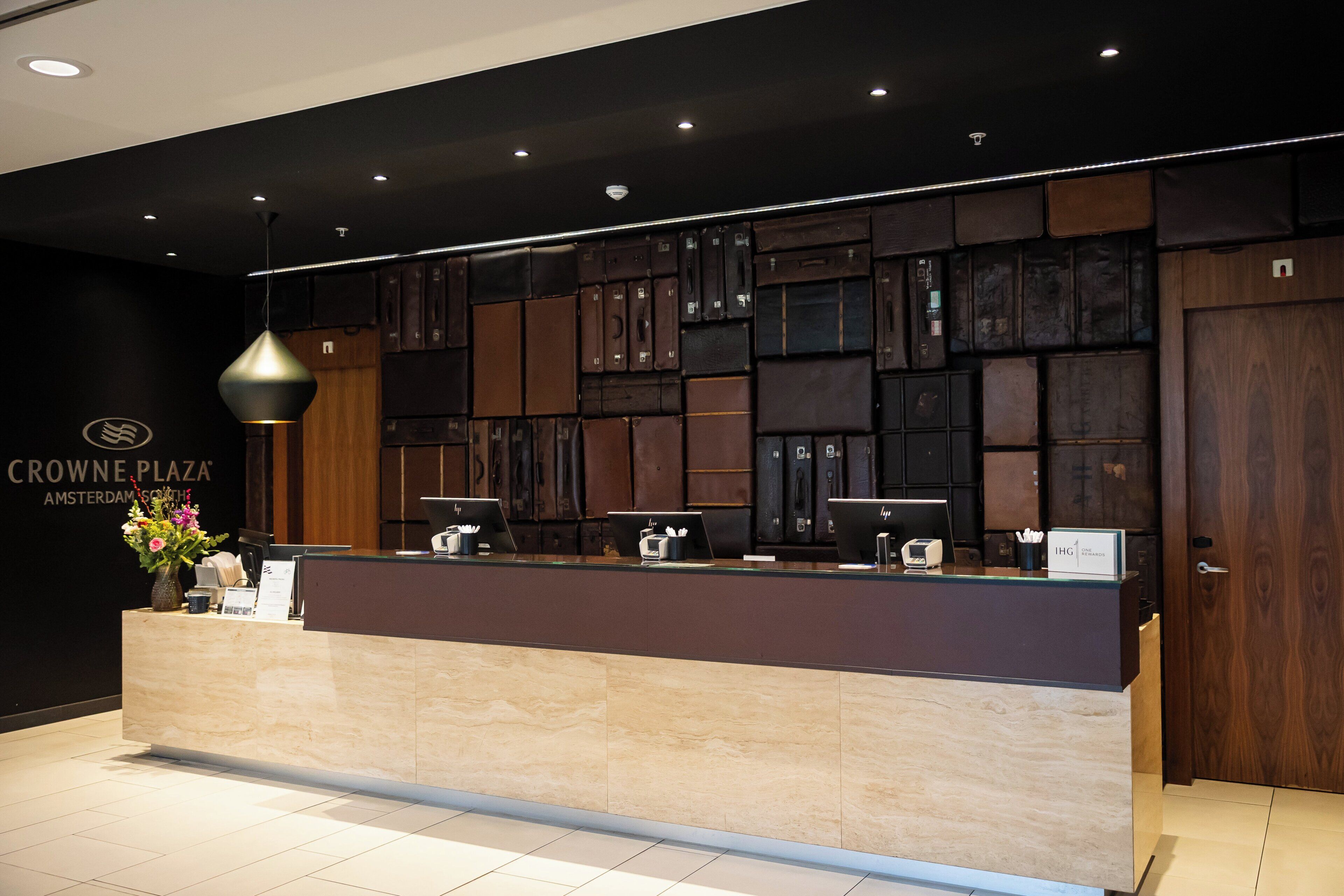A reception desk with a dark wood panel backdrop, a modern pendant light, and a sign for Crowne Plaza Amsterdam.