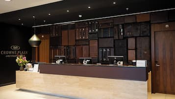 A reception desk with a dark wood panel backdrop, a modern pendant light, and a sign for Crowne Plaza Amsterdam.