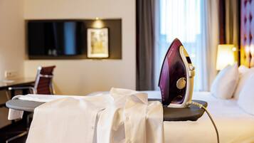 A hotel room with a bed, an ironing board with an iron, a desk, a chair, and a window with curtains.