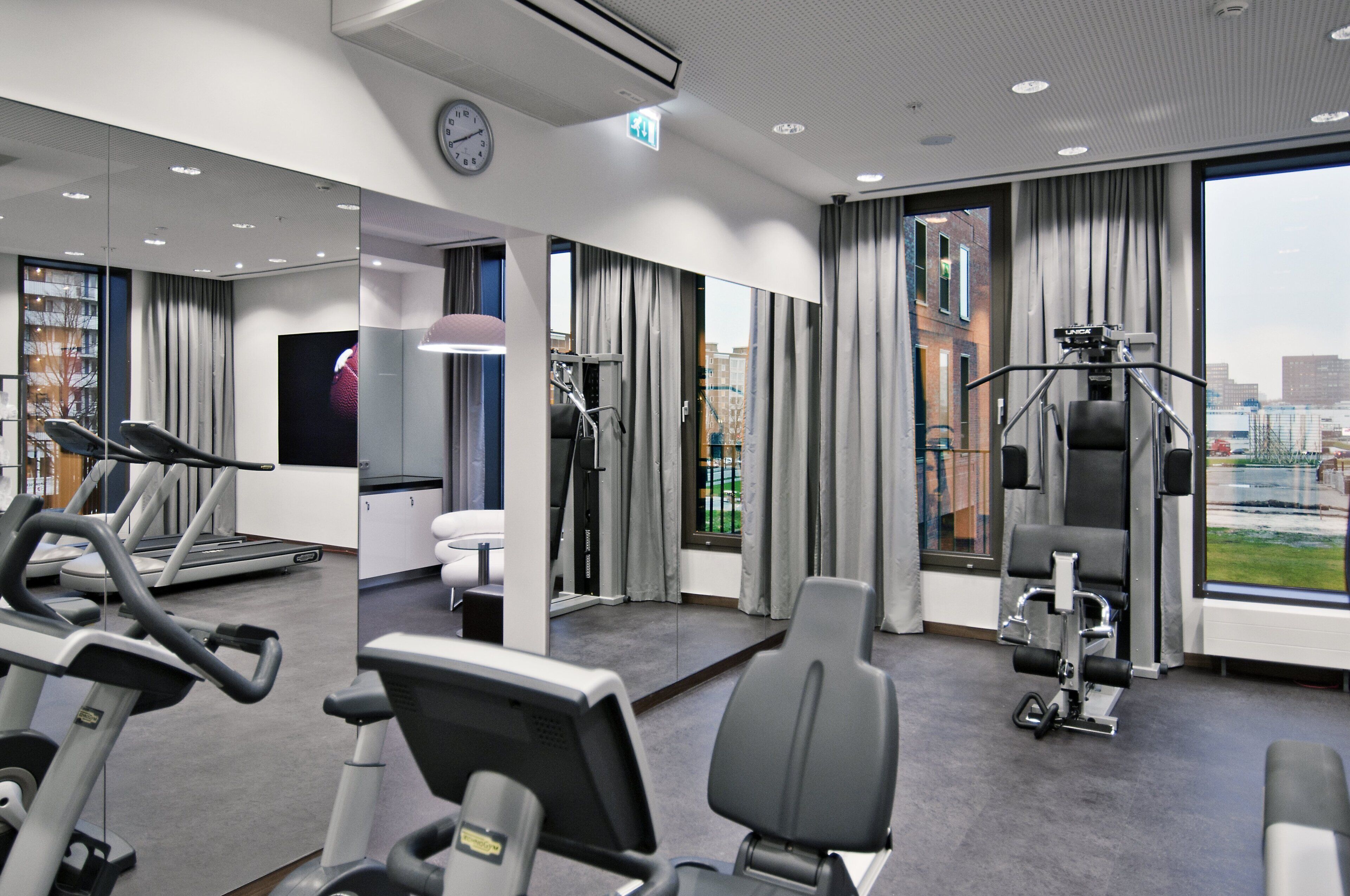 A gym with exercise equipment, a clock, and a view of buildings through large windows.