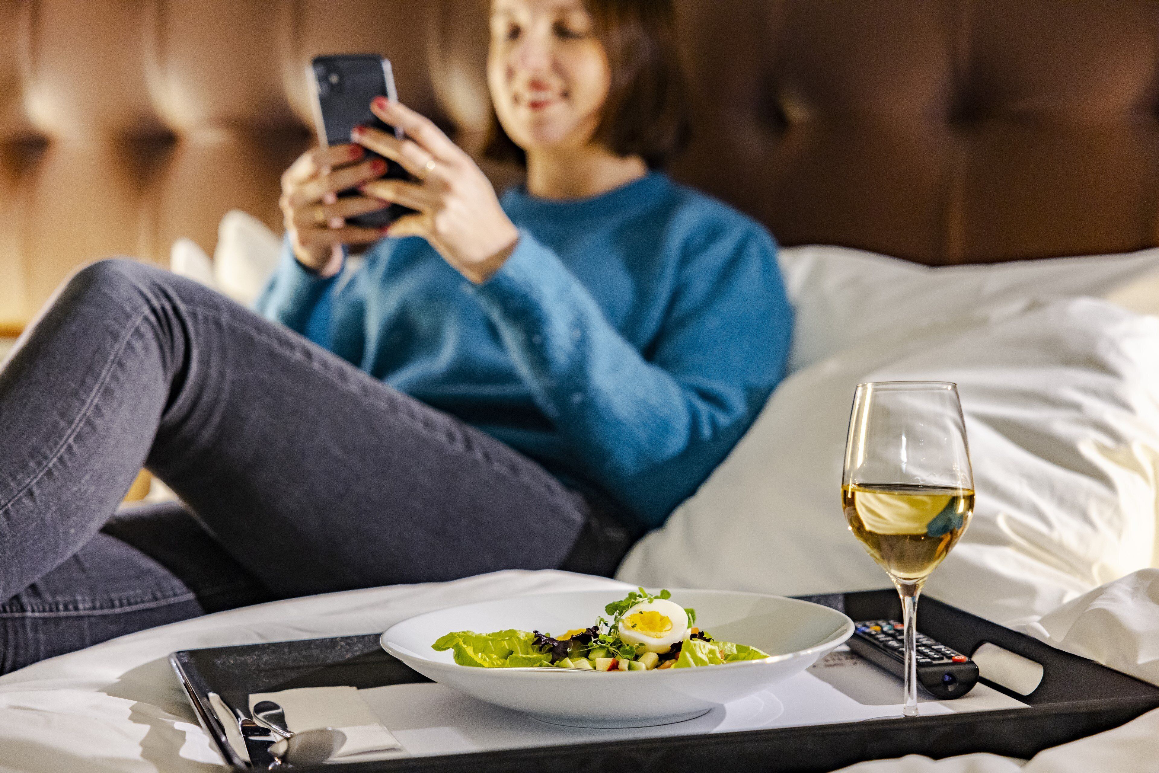 Una persona recostada en una cama con un control remoto, una copa de vino blanco y un plato de comida.
