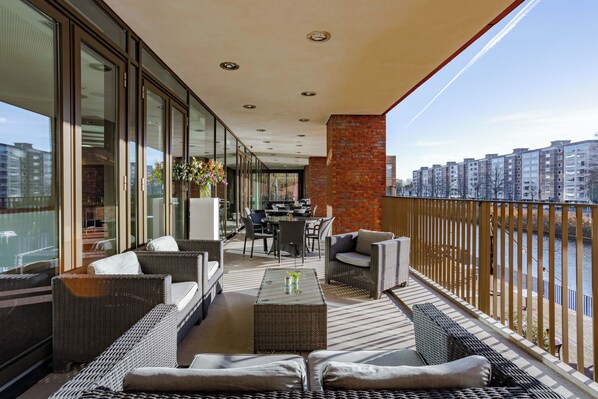Un balcón con mobiliario de mimbre, una mesa con flores y vistas a edificios y un río.