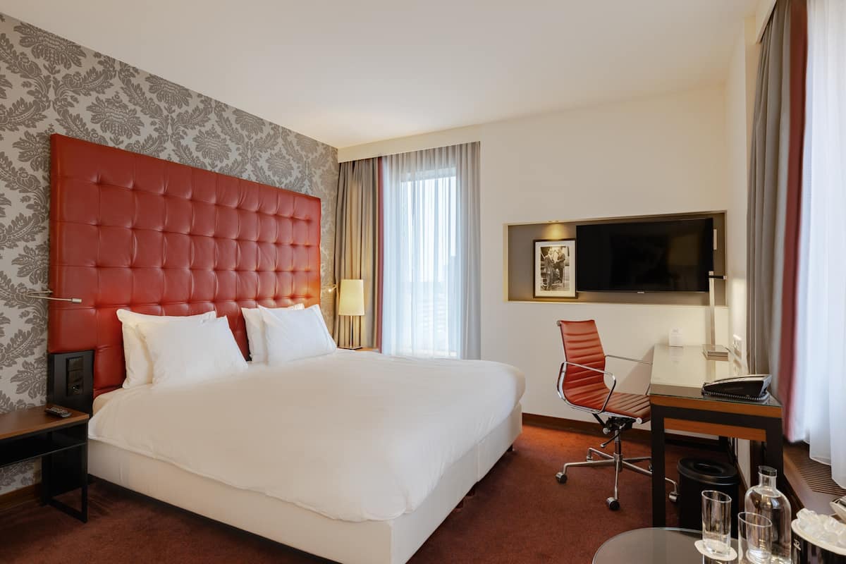 Une chambre d’hôtel avec un grand lit, un bureau, une chaise, une télévision et une fenêtre avec des rideaux.