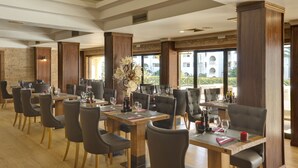A restaurant with multiple tables and chairs, wooden flooring, and large windows offering a view of the outdoors. - Paladim & Alagoamar Hotels (Albufeira)