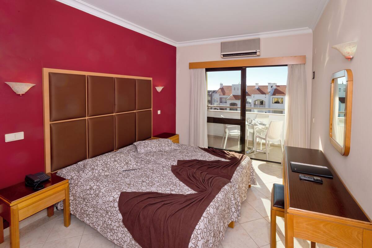 Habitación de hotel con una cama grande, un escritorio, una silla, una mesita de noche y vistas a la ciudad a través de un ventanal.