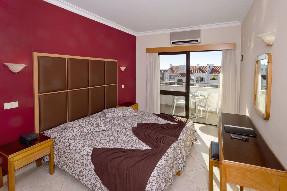 Habitación de hotel con una cama grande, un escritorio, una silla, una mesita de noche y vistas a la ciudad a través de un ventanal.