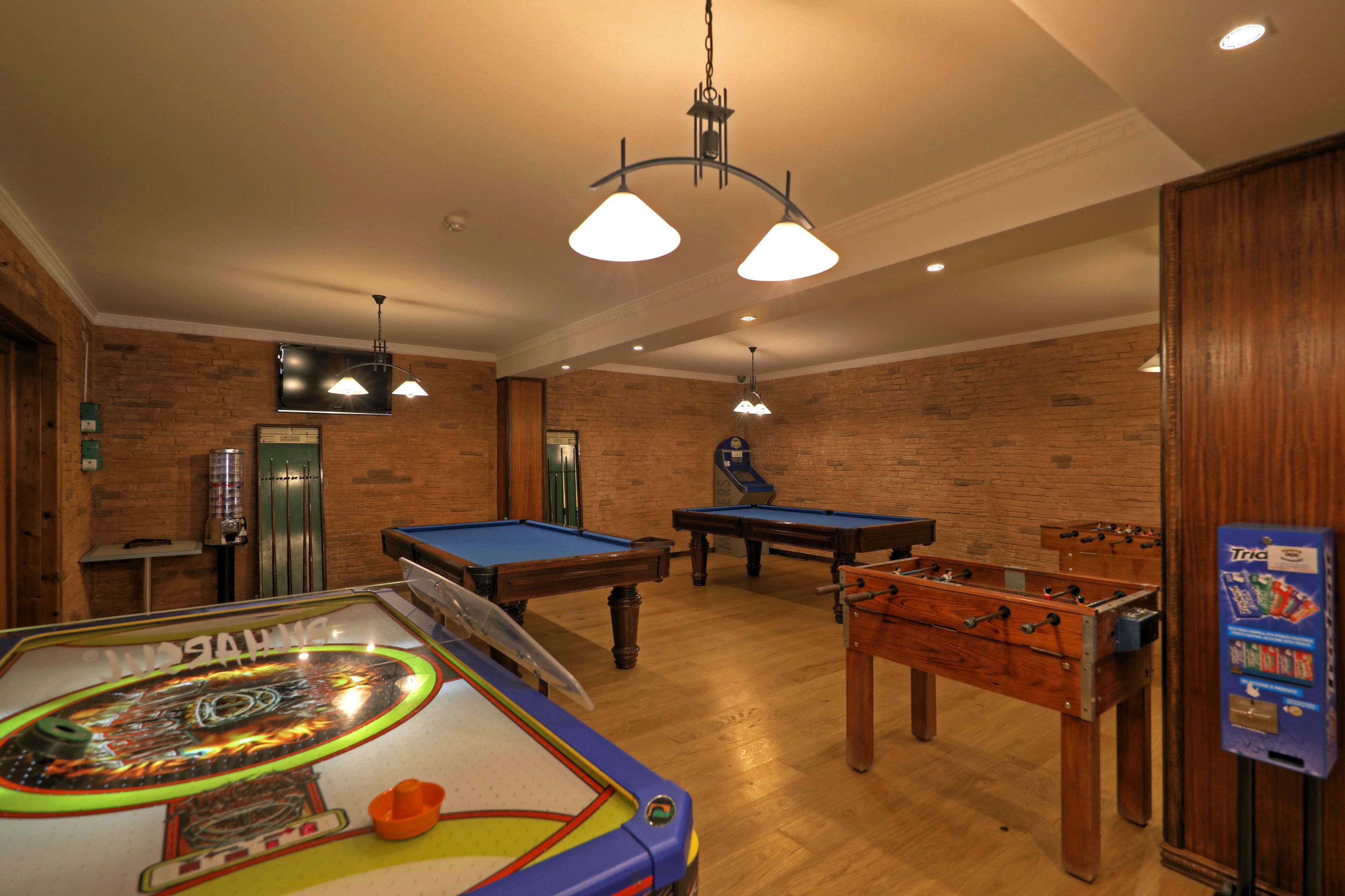 A room with a foosball table, pool table, and a ping pong table. There are ceiling lights and a brick wall.