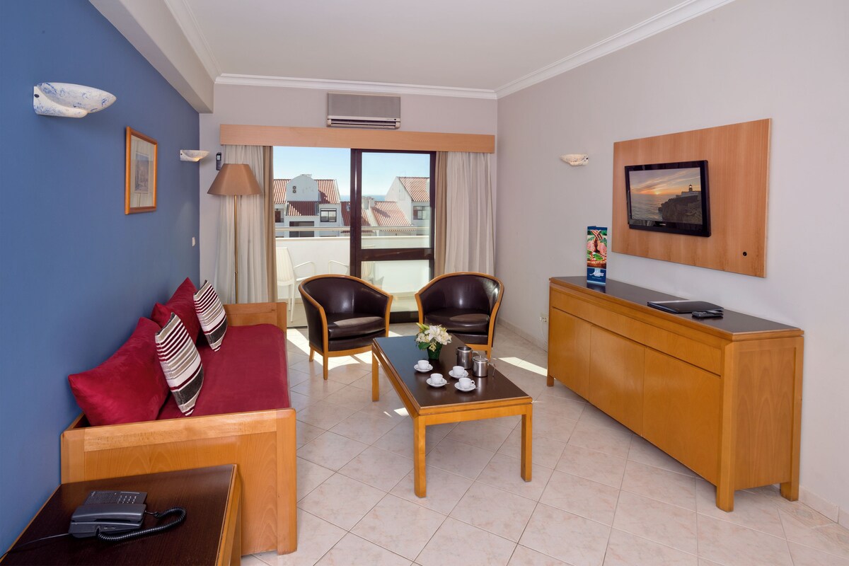 Hotelkamer met een bank, twee stoelen, een salontafel, een bureau, een flatscreen-tv en een groot raam met uitzicht op gebouwen.