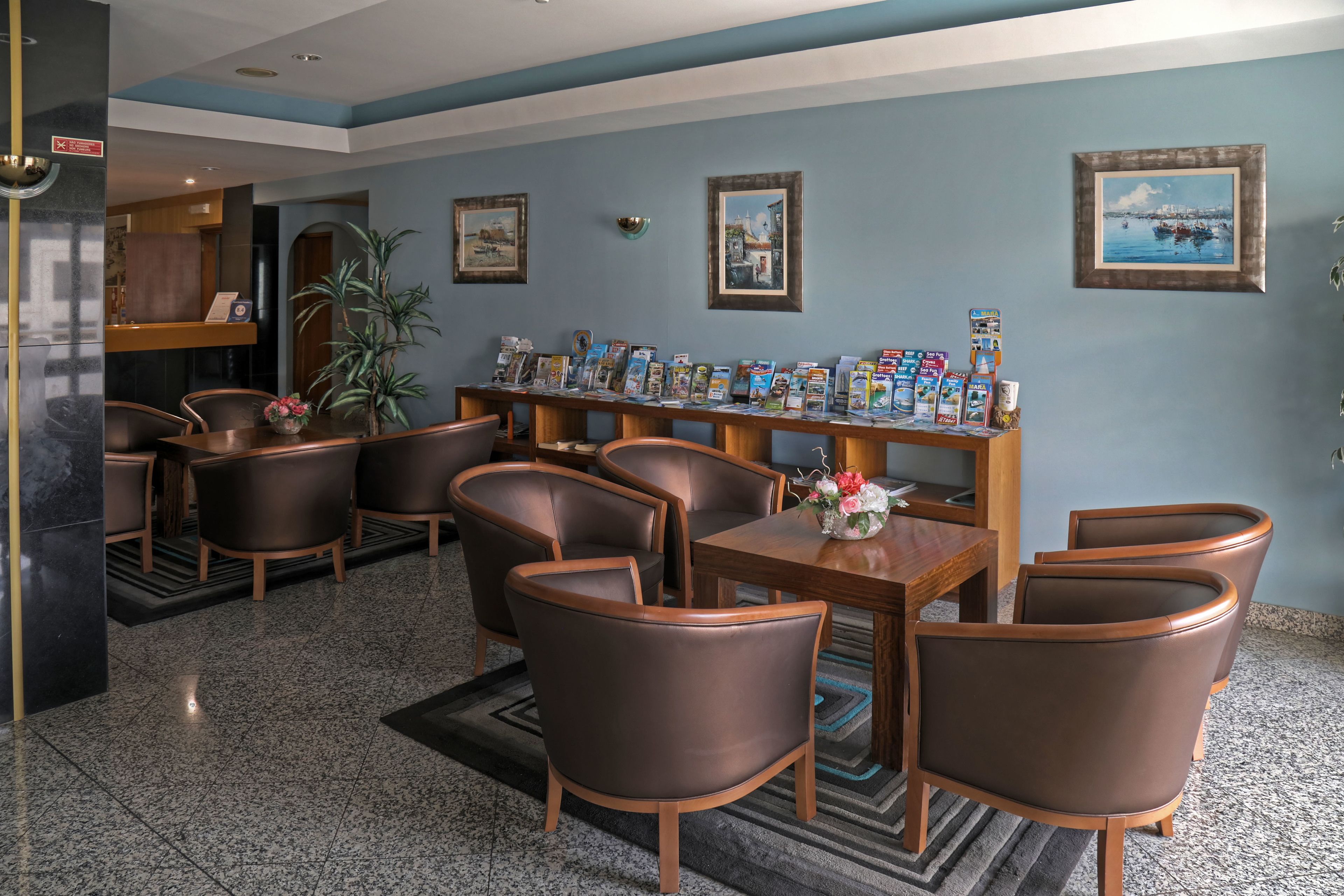 Un hall con mostrador de recepción, zona de estar con sillas y una mesa, cuadros en la pared y una exhibición de folletos.
