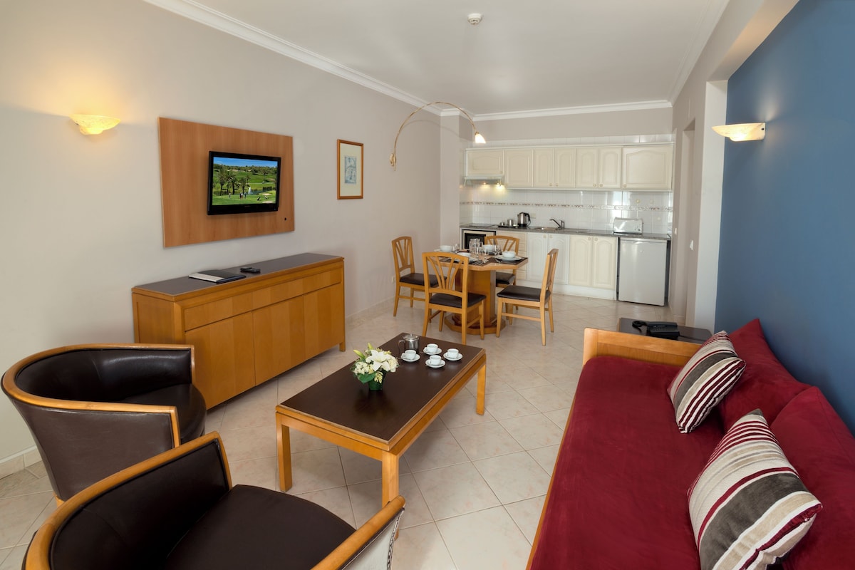 A modern living room with a wooden cabinet, a TV mounted on the wall, a dining area with a table and chairs, and a kitchen in the background.
