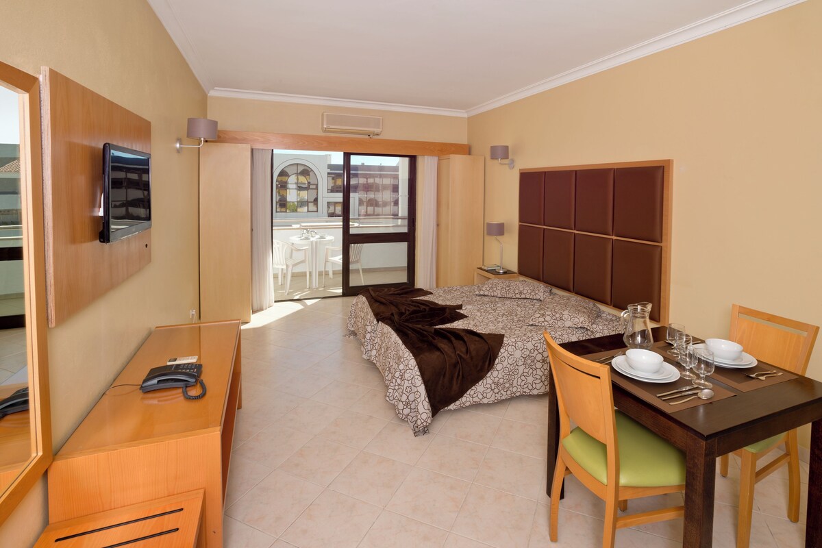 A hotel room with a bed, a desk with a telephone, a dining table set for two, and a view of the outside balcony.