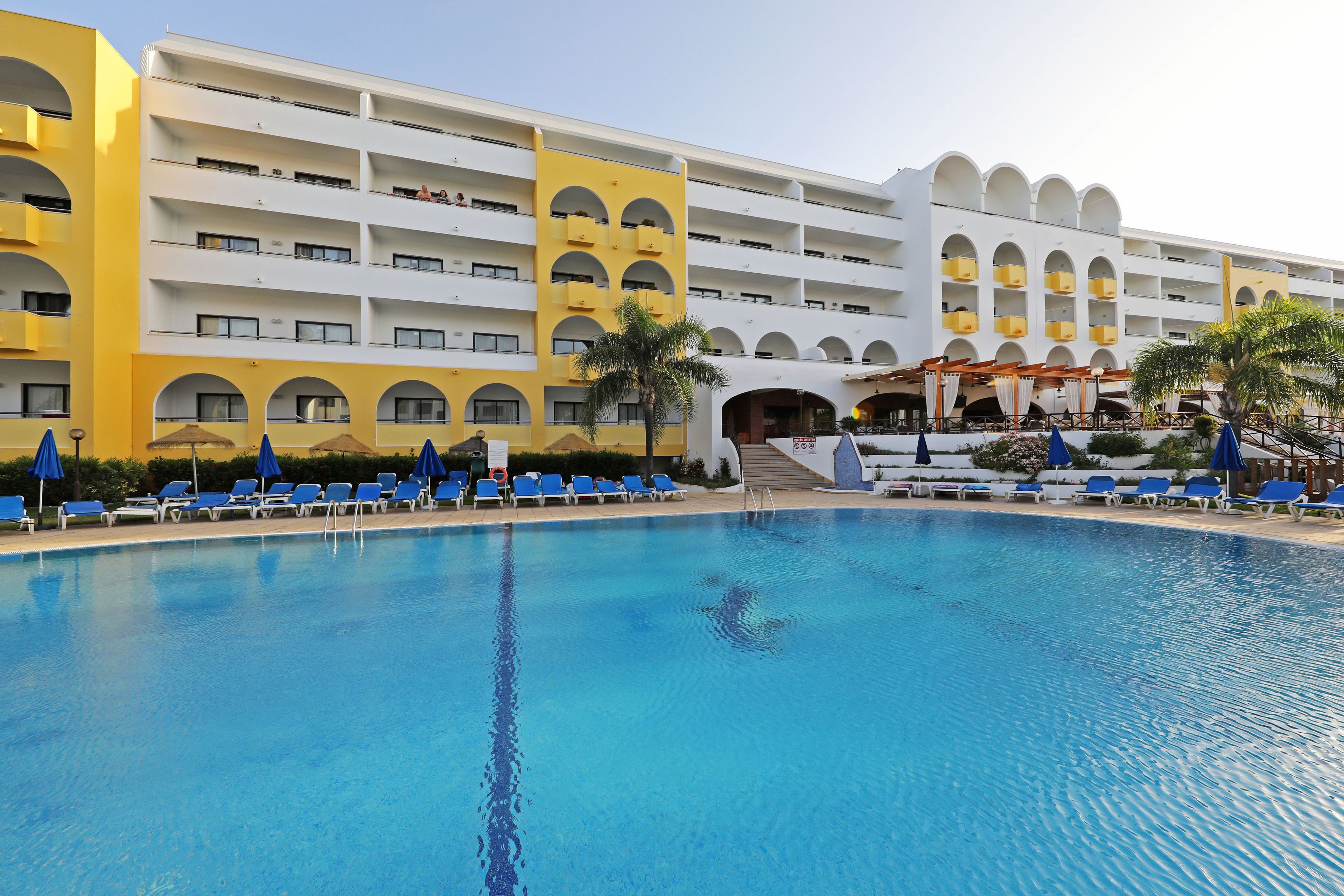 A hotel with a large swimming pool, multiple balconies, and blue lounge chairs.