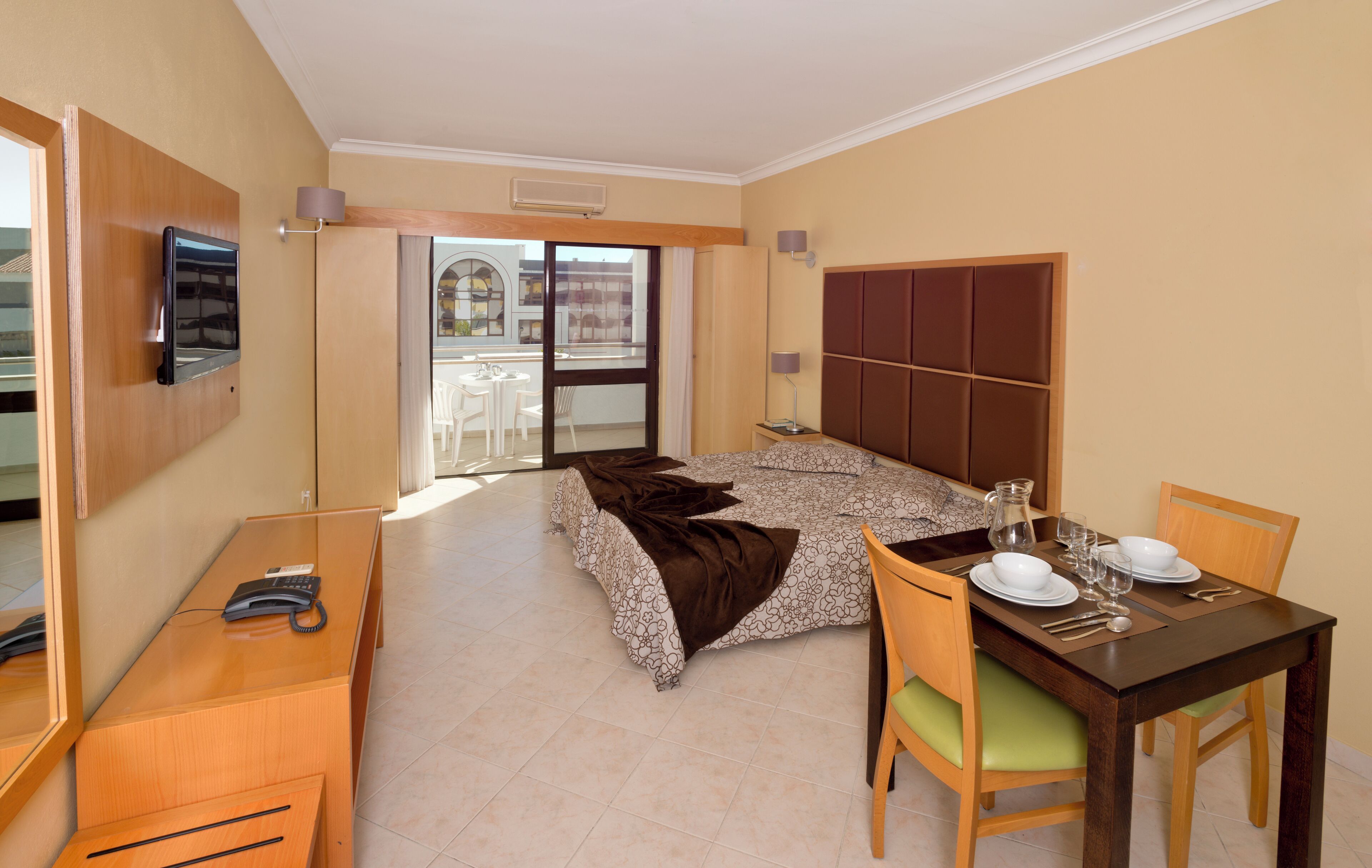 Une chambre d’hôtel avec un lit, un bureau avec un téléphone, une table à manger dressée pour deux personnes et une vue sur le balcon extérieur.