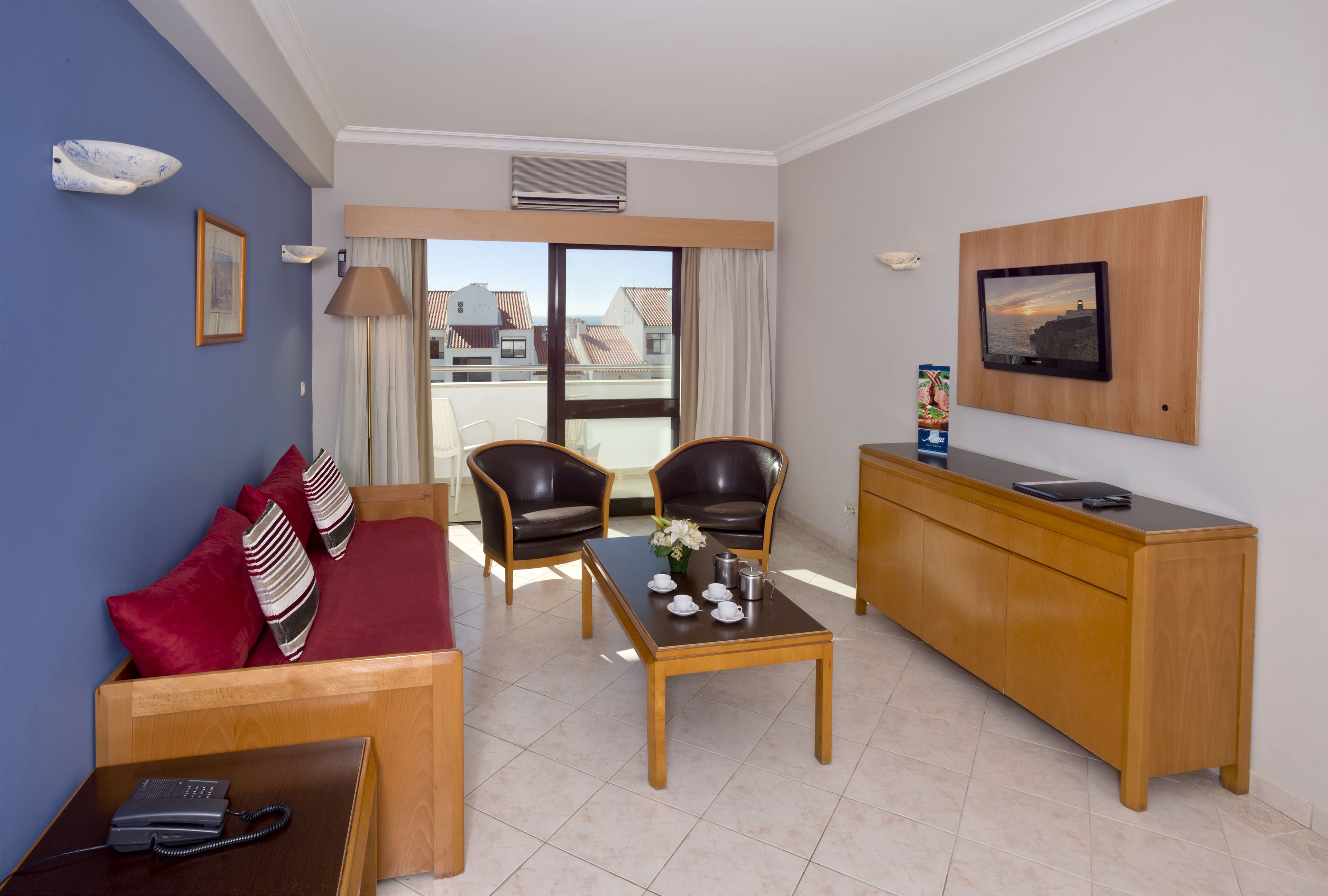 Habitación de hotel con sofá, dos sillones, una mesa de centro, un escritorio, un televisor de pantalla plana y un ventanal con vistas a edificios.