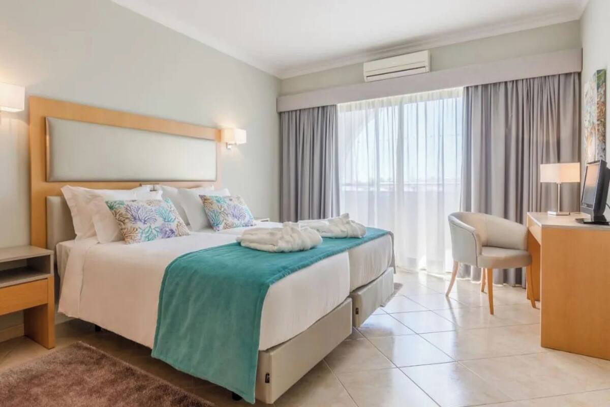 A hotel room with a bed, bedside tables, a desk, a chair, a TV, and a window with curtains.