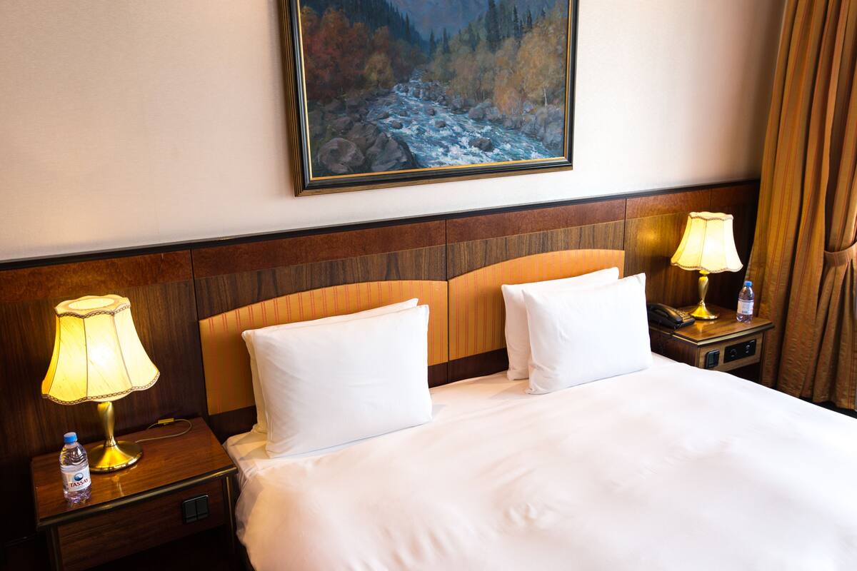 A hotel room with a large bed, two bedside lamps, a painting of a river, and a wooden headboard.