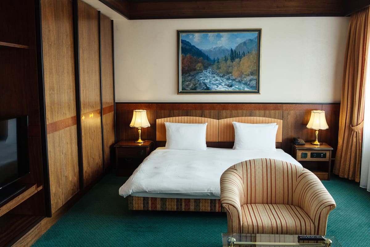 A hotel room with a large bed, two bedside tables with lamps, a television, a chair, and a painting of a mountainous landscape on the wall.