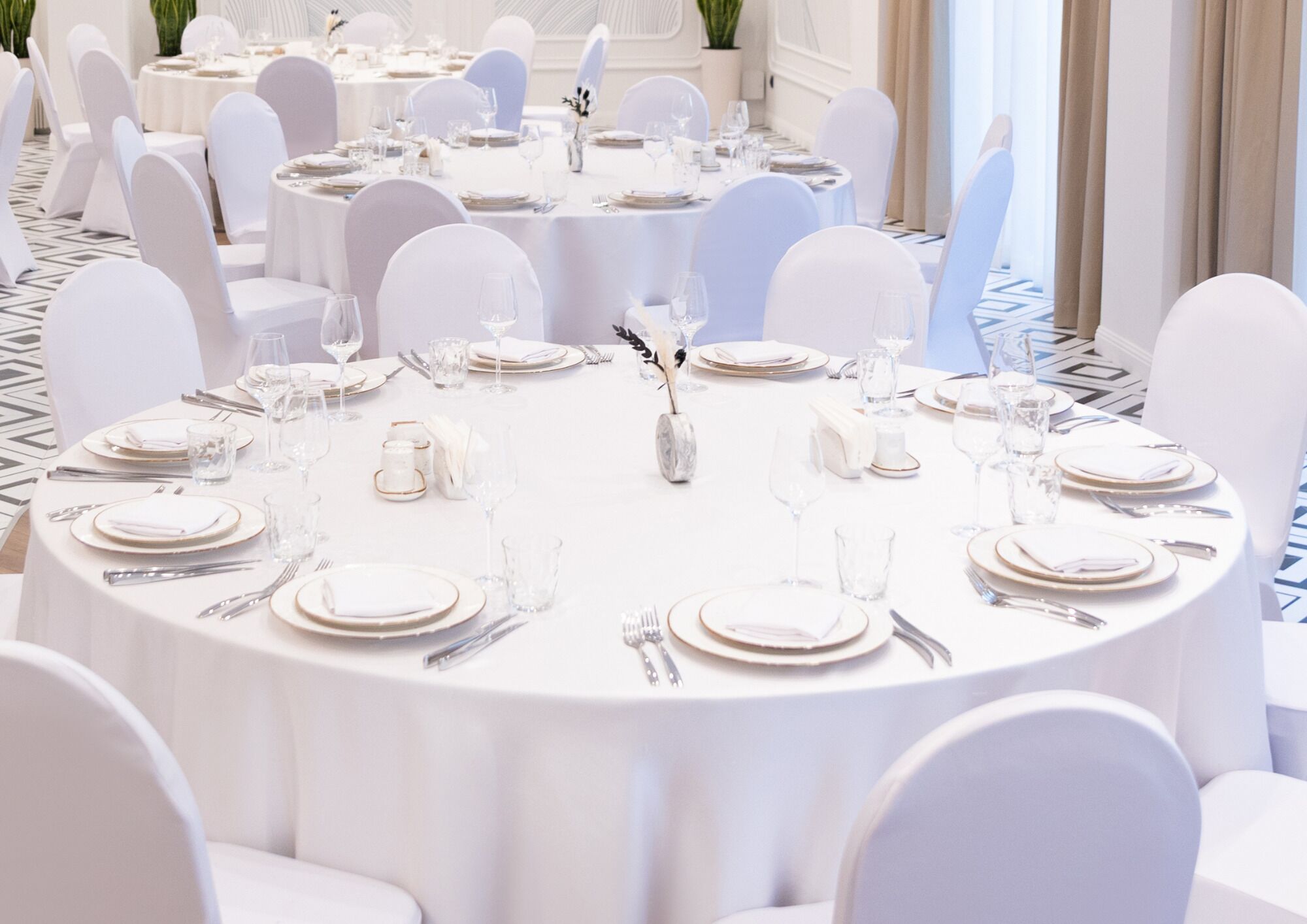 A banquet hall with round tables set for dining, white chairs, and a patterned floor.