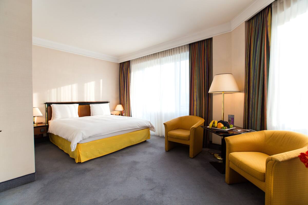 A hotel room with a bed, two chairs, a table with fruit, and a lamp.