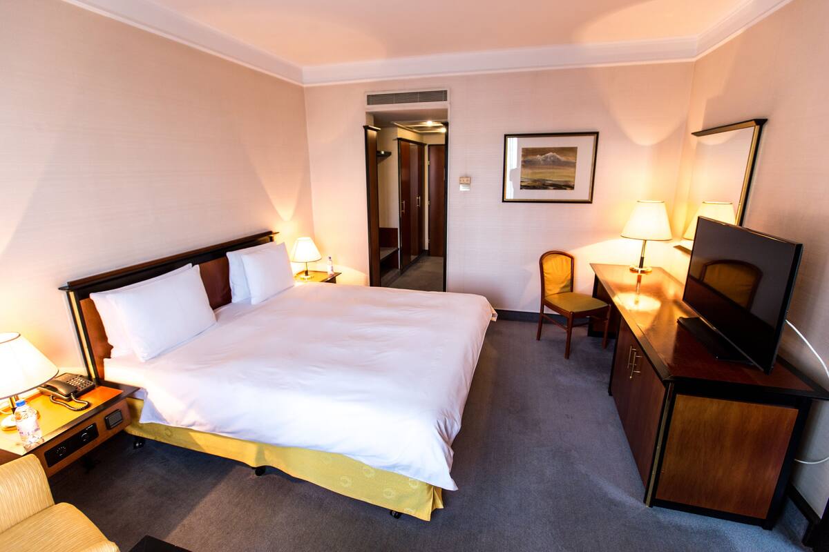 A hotel room with a bed, bedside table, lamp, chair, desk, TV, and a painting on the wall.