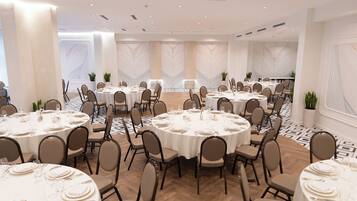 A large conference room with multiple round tables, each set with white tablecloths, plates, and cutlery.